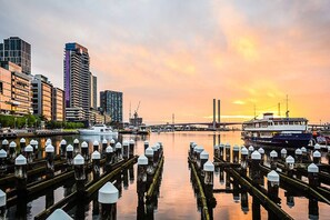 A waterfront cityscape with modern buildings, a dock with moored boats, and a sunset in the background. - Lovely Docklands 2 Bedroom (Docklands)