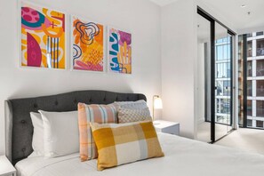 Een moderne slaapkamer met een grijs gestoffeerd hoofdbord, een wit sprei en diverse gestreepte kussens. Boven het bed hangen abstracte schilderijen aan de muur.