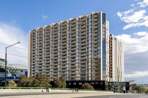 A high-rise residential building with multiple balconies, a pedestrian walkway, and a bridge in the background. - Modern 2 Bedroom with Parking (Docklands)