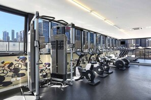 Un gimnasio con diversos equipos de ejercicio, amplias ventanas y vistas a la ciudad.