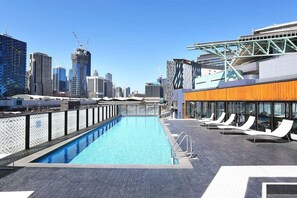 Una piscina en la azotea con un horizonte urbano de fondo, sillas de descanso y un edificio moderno con fachada de vidrio.