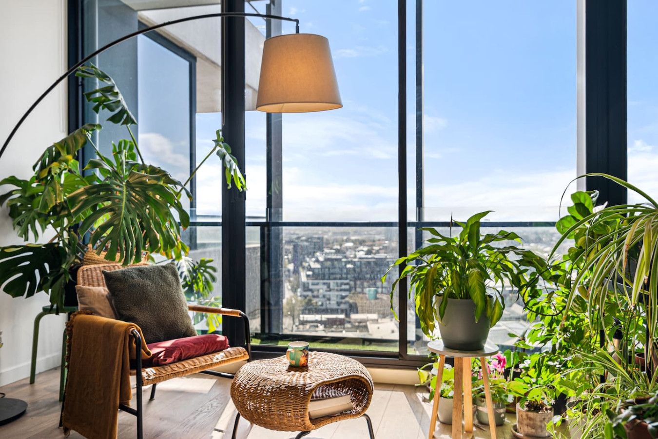A high-rise apartment with a large window, a wicker chair, a floor lamp, and various potted plants.