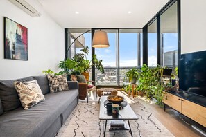 Een moderne woonkamer met een grijze bank, een salontafel en een televisie. Er staan verschillende planten en een groot raam met uitzicht op de stad.