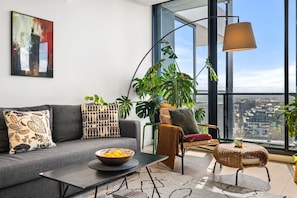 A modern living room with a grey sofa, a coffee table, and a wicker chair. There is a large window with a view of the city. - Green 1 Bedroom by Flagstaff (West Melbourne)