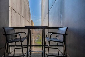 Um corredor externo estreito com grade de metal, duas cadeiras de metal preto e uma mesa.