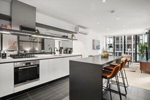 A modern kitchen with a central island, bar stools, and a view of the city through large windows. - Docklands 2 Bedroom with Pool (Docklands)