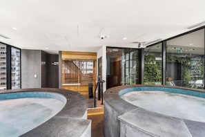 A modern indoor swimming pool area with two hot tubs, a wooden staircase, and a glass wall with a view of greenery. - Central 2 Bedroom with Sauna (Melbourne)