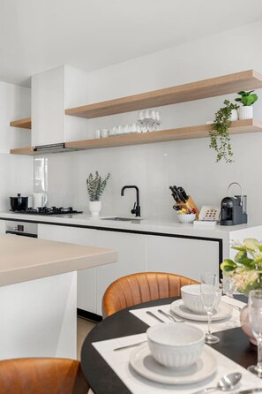 A modern kitchen with white cabinetry, wooden shelves, a black faucet, and a coffee machine. - Chic 2 Bedroom near Marvel (Docklands)