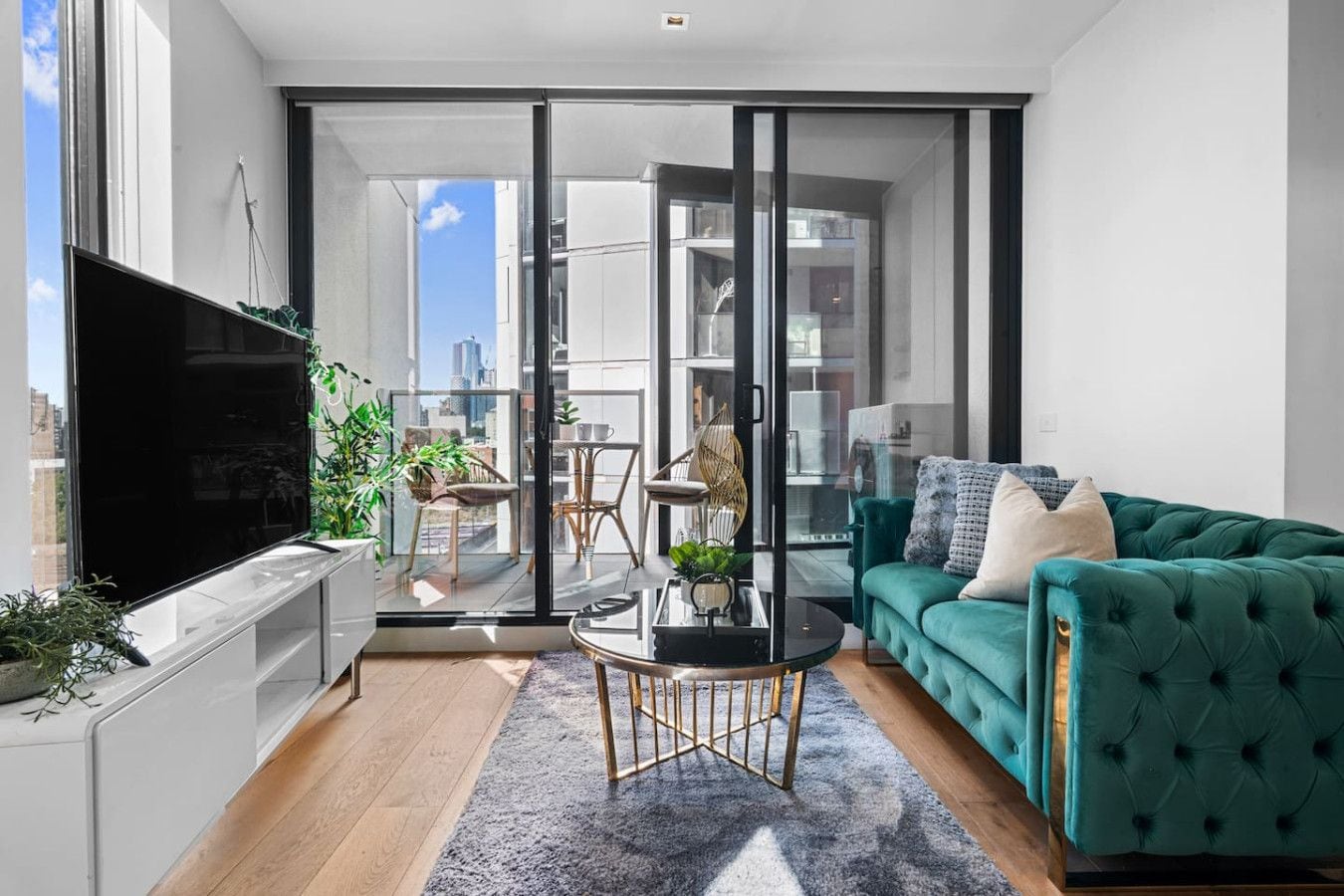 A modern living room with a flat-screen TV, a teal sofa, a coffee table, and a view through a sliding glass door to an outdoor balcony.