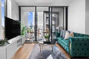 A modern living room with a flat-screen TV, a teal sofa, a coffee table, and a view through a sliding glass door to an outdoor balcony. - Gorgeous 2 Bedroom by Marvel (Docklands)