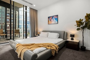 A modern hotel room with a large bed, a view through floor-to-ceiling windows, a painting on the wall, and a potted plant. - Luxury Penthouse by Marvel (Docklands)