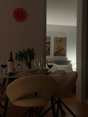 Un coin repas avec une table dressée pour deux personnes, des verres à vin et une bouteille de vin. Il y a une horloge au mur et une plante sur la table.