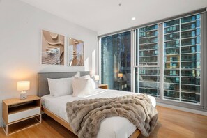 A modern bedroom with a large bed, a nightstand, a lamp, and a view of a cityscape through floor-to-ceiling windows. - Skyline Oasis 3 Bedroom (Melbourne)