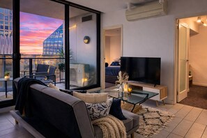 Un saló modern amb un sofà, una taula de centre i una televisió. Hi ha un balcó amb vista a un paisatge urbà al capvespre.