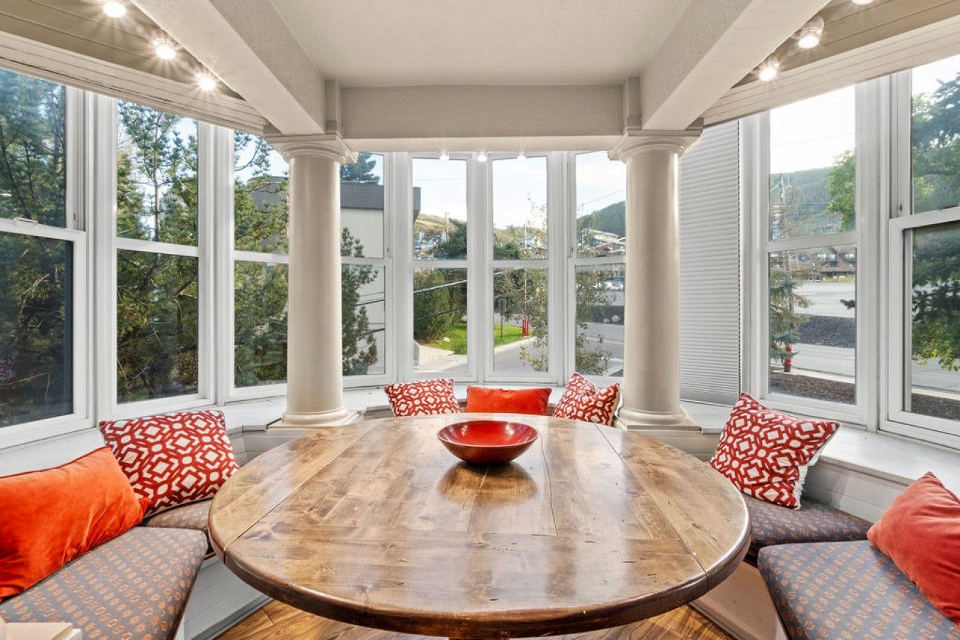 A dining area with a round wooden table, red and patterned cushions, and large windows offering a view of a residential street.