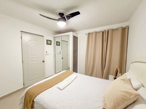 A bedroom with a bed, a ceiling fan, a door, and a window with curtains. - Secure and Central 2BR Apt (Sosúa)