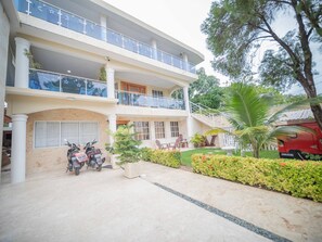 A multi-story building with balconies, a garden area, and parked scooters. - Secure and Central 2BR Apt (Sosúa)