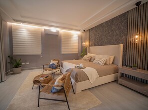 A modern bedroom with a large bed, a seating area with chairs, a bedside table, and a decorative wall panel. - Stylish Luxury Villa in Sosua- 5BR Suites! (Sosúa)