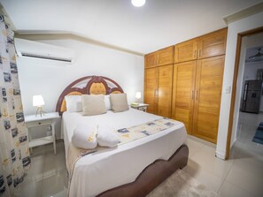 Quarto com cama, mesas de cabeceira, ar condicionado, guarda-roupa de madeira e banheiro visível por uma porta aberta.