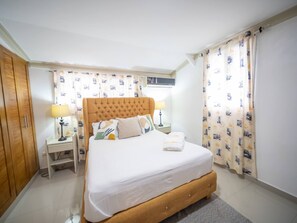 A bedroom with a large bed, wooden wardrobe, bedside lamps, and patterned curtains. - Modern  2BR Apt Central in Sosua (Sosúa)
