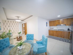 A modern living room with a glass coffee table, blue armchairs, and a dining area with a glass table. - Modern  2BR Apt Central in Sosua (Sosúa)