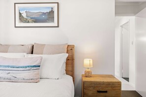 A bedroom with a wooden headboard, a bed with white bedding, a nightstand with a lamp, and a framed picture on the wall. - Saltwater Haven - Pool, Beach & Dog-Friendly (Mooloolaba Region)