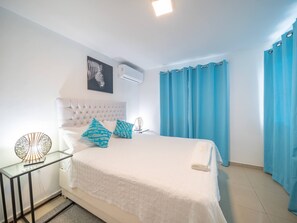 A bedroom with a bed, a bedside table, a wall-mounted air conditioner, and a window with curtains. - Cozy & Central 3BR Apt Secure & Comfortable (Sosúa)