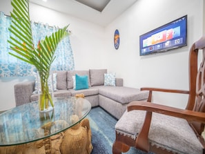 A modern living room with a sectional sofa, a glass coffee table, a wooden chair, a flat-screen TV mounted on the wall, and a tropical plant. - Cozy & Quiet 1BT Apt Central in Sosua (Sosúa)