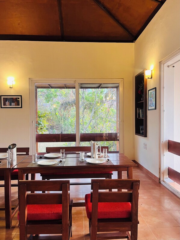 A dining area with a wooden table set for four, surrounded by red cushioned chairs, large windows with a view of trees, and wall-mounted light fixtures.