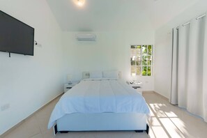 Une chambre à coucher comprenant un lit, un téléviseur fixé au mur, un appareil de climatisation, une table de chevet avec une lampe, une fenêtre avec des rideaux et une vue sur la verdure à l’extérieur.