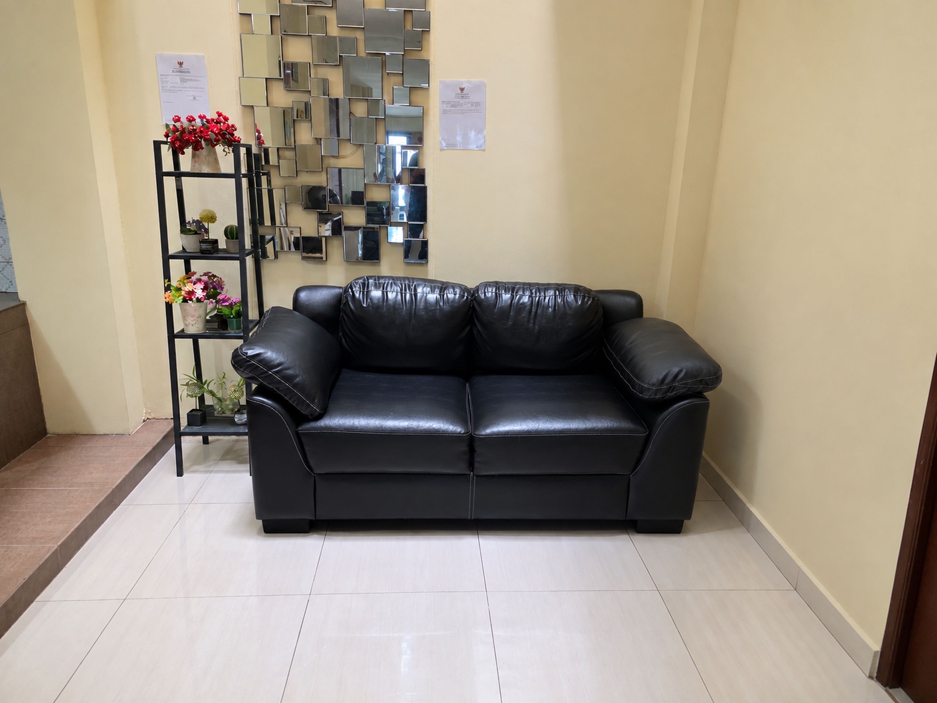 A black leather sofa in a room with a tiled floor and a wall decorated with mirrors and a small shelf with flowers.