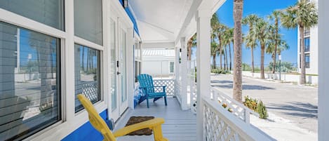 A covered porch with two chairs, one blue and one yellow, overlooking a beach with palm trees.