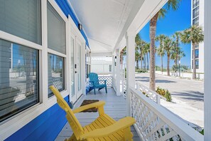 A covered porch with two chairs, one blue and one yellow, overlooking a beach with palm trees. - Blue Lagoon at White Sands| 2br/1ba close to beach (Indian Rocks Beach)