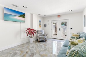 A lobby with a flat-screen TV, a potted plant, a patterned armchair, a couch with patterned pillows, and a door with an exit sign. - White Sands Beach Cottages| 6 cottages sleeps 32 (Indian Rocks Beach)