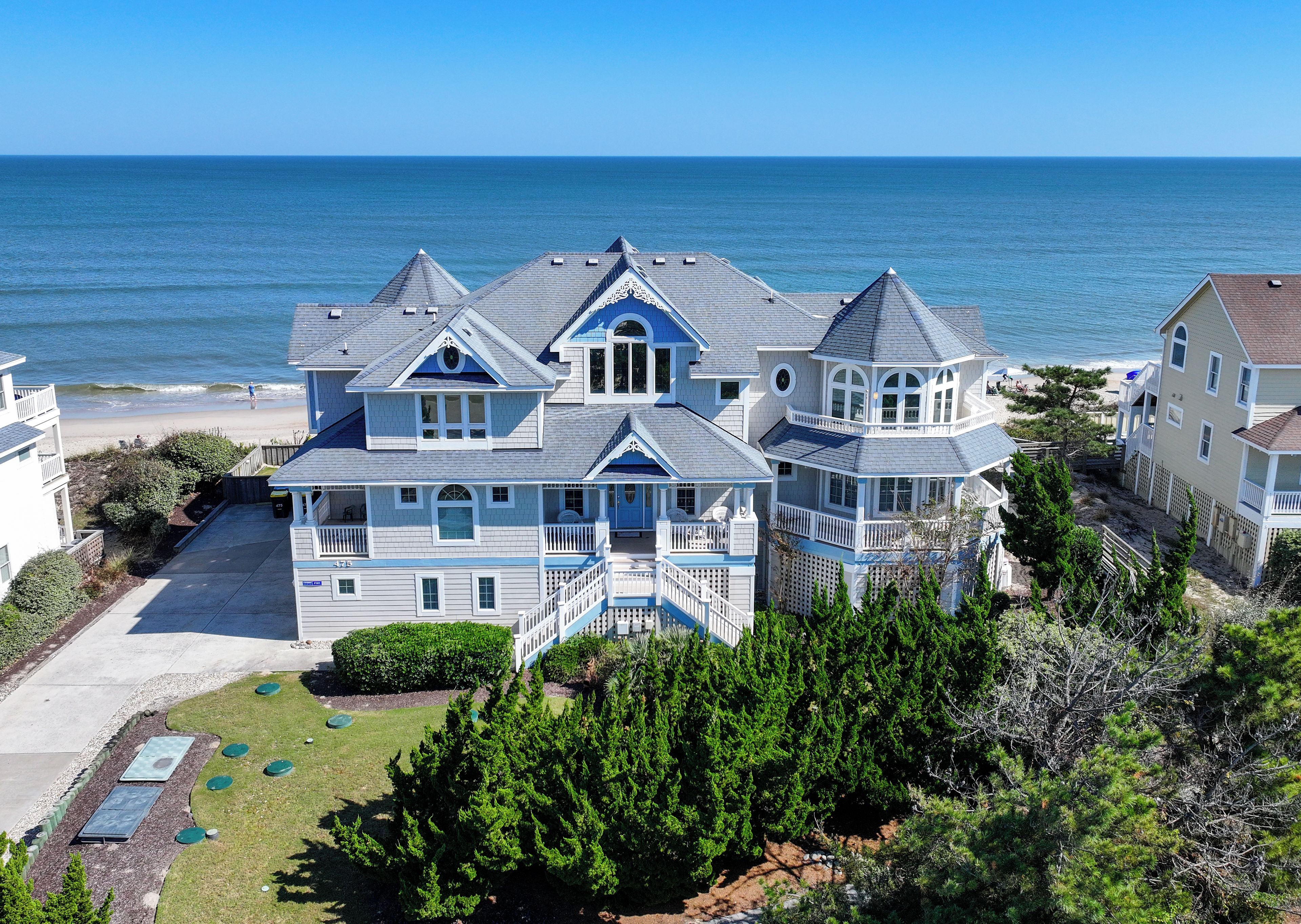 A large, multi-story house with a balcony overlooking the ocean, a driveway, and a well-maintained lawn.