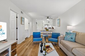 Ein modernes Wohnzimmer mit Fernseher, zwei blauen Sesseln, einem beigen Sofa und einem Essbereich mit Tisch und Stühlen.