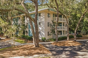 A multi-story building with a light-colored exterior, surrounded by mature trees and a well-maintained lawn. - Fiddler's Cove Villa with Balcony, Tennis, Pickleball & Heated Community Pool (Hilton Head Island)