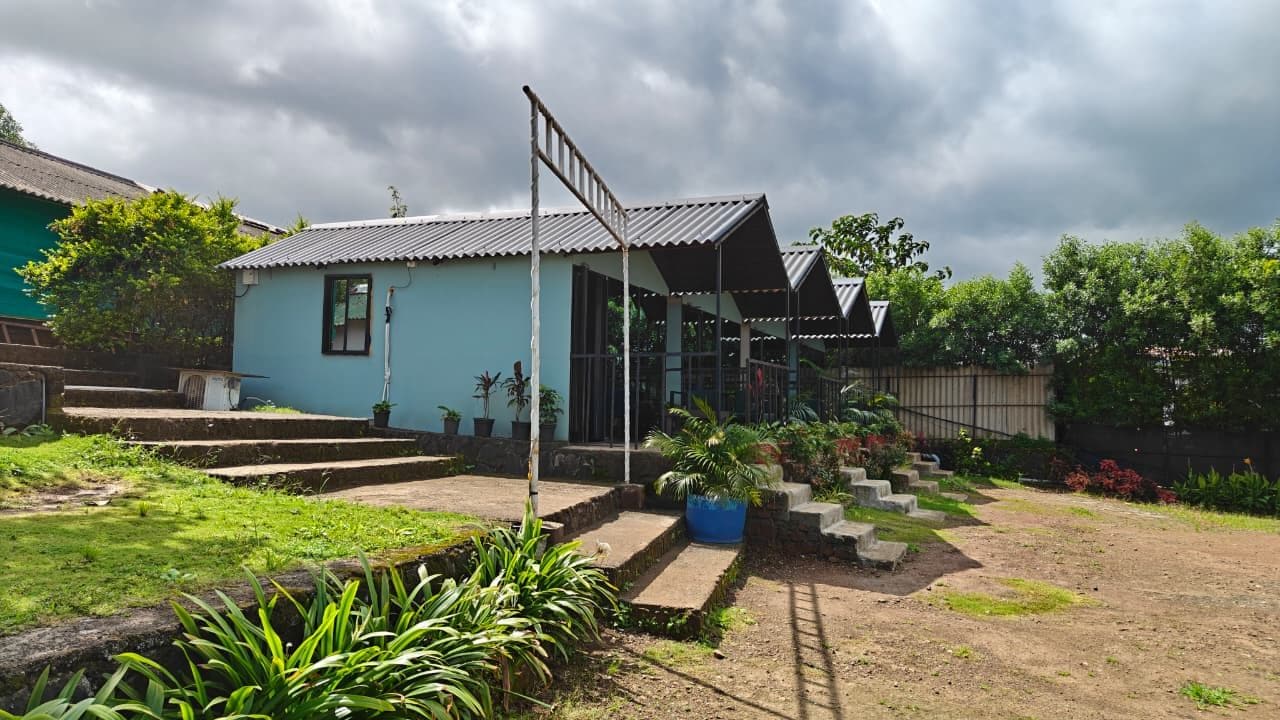 Um edifício térreo com telhado inclinado, varanda com degraus até a entrada e área de jardim com diversas plantas e um pequeno elemento aquático.