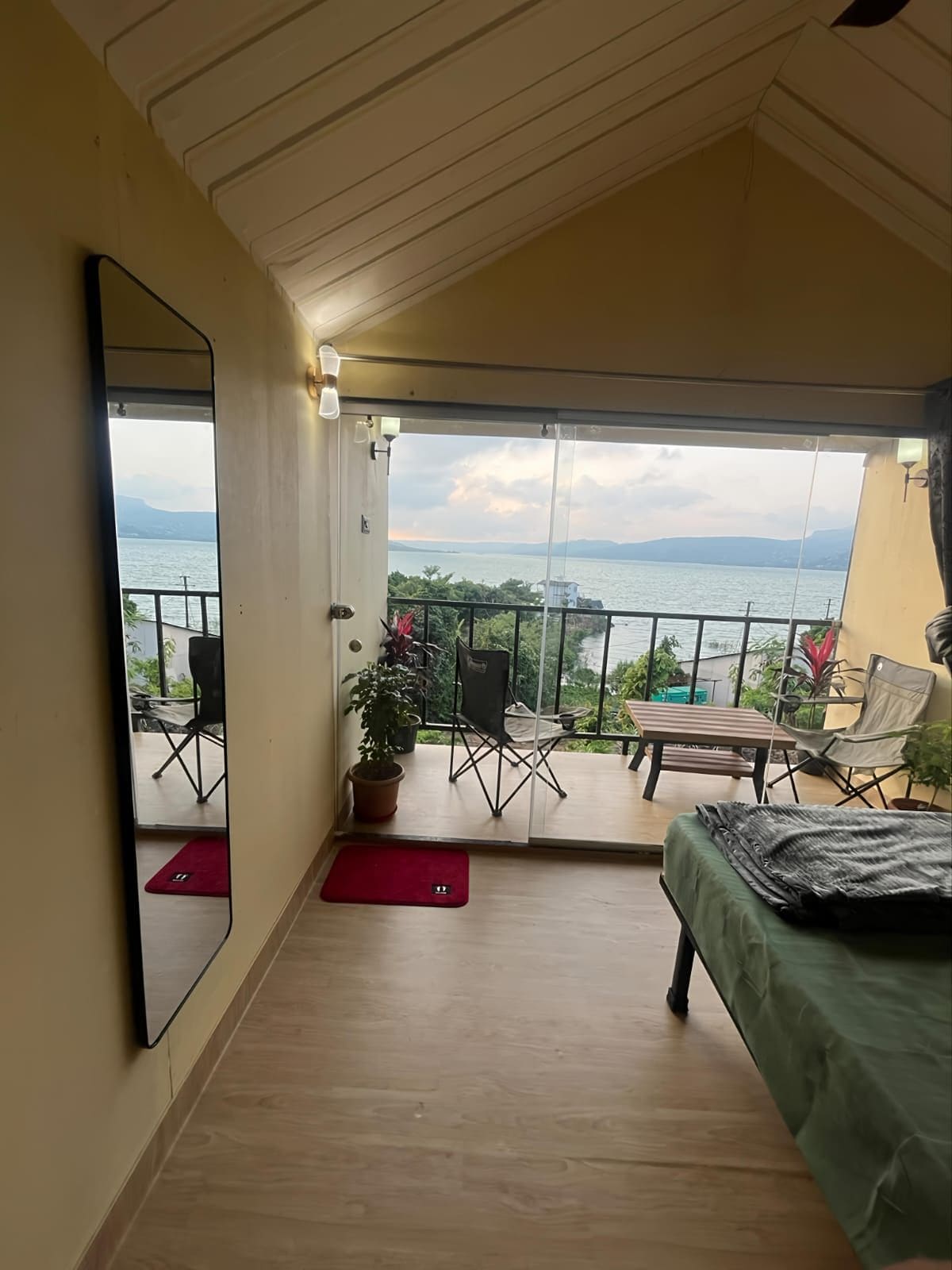 A room with a bed, a mirror, a balcony with a view of the sea, and a potted plant.