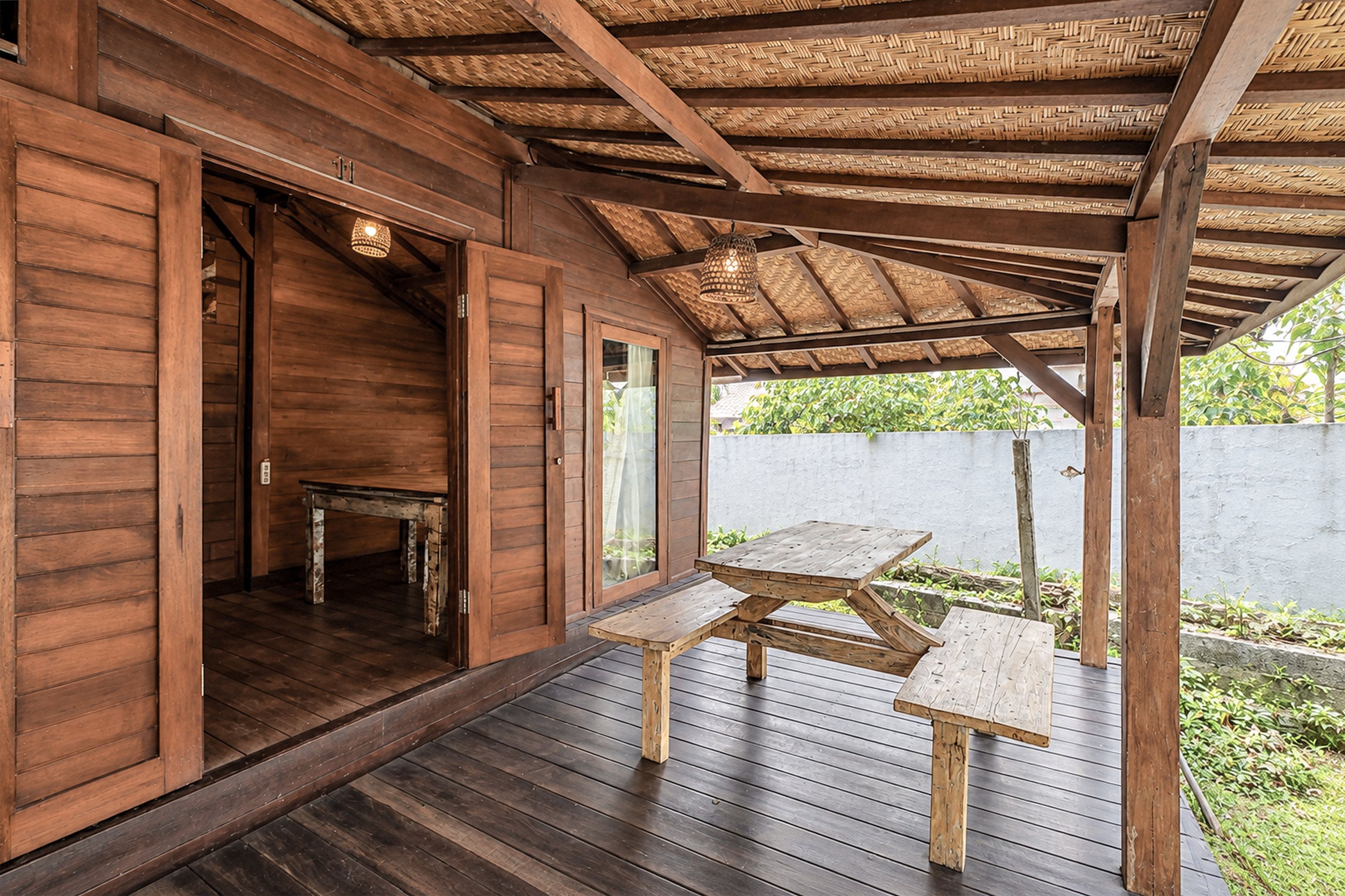 Een houten terras met een tafel en stoelen, een overdekt gedeelte met een geweven plafond, en uitzicht op groen.