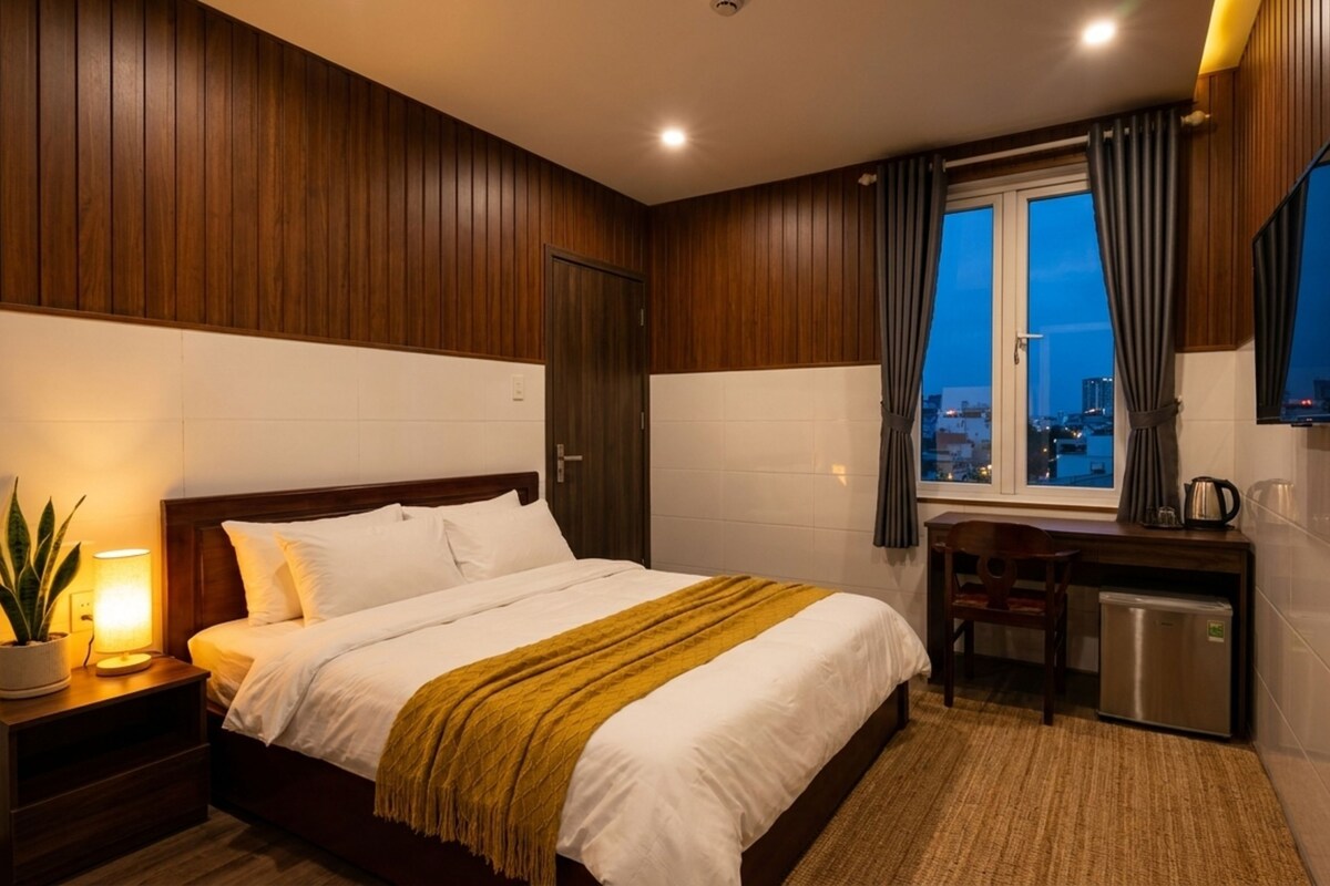Een hotelkamer met een bed, nachtkastje, lamp, bureau, stoel en een raam met uitzicht over de stad.
