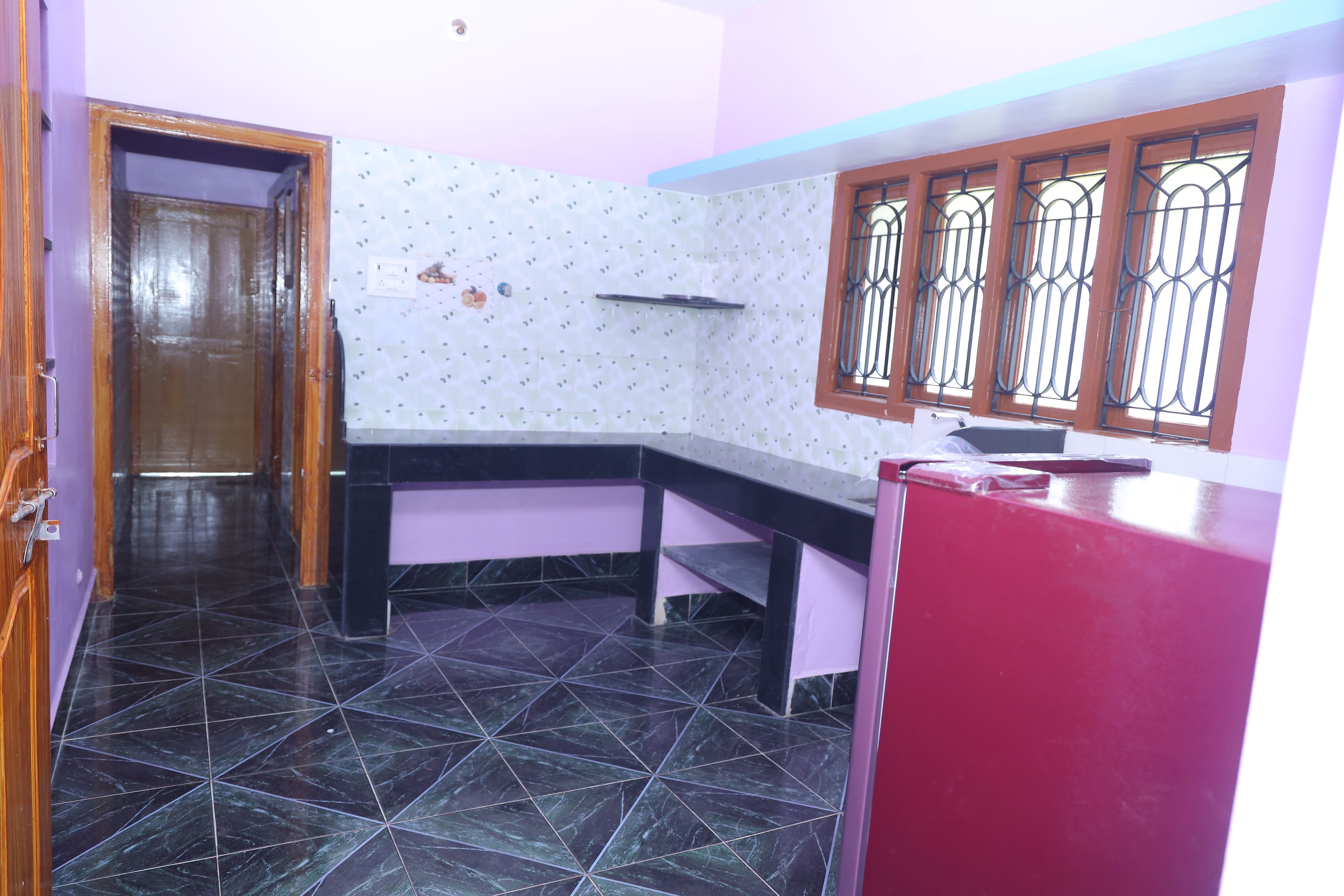 Una cocina con piso de baldosas negras, un refrigerador rojo, una puerta de madera y una ventana con rejas.