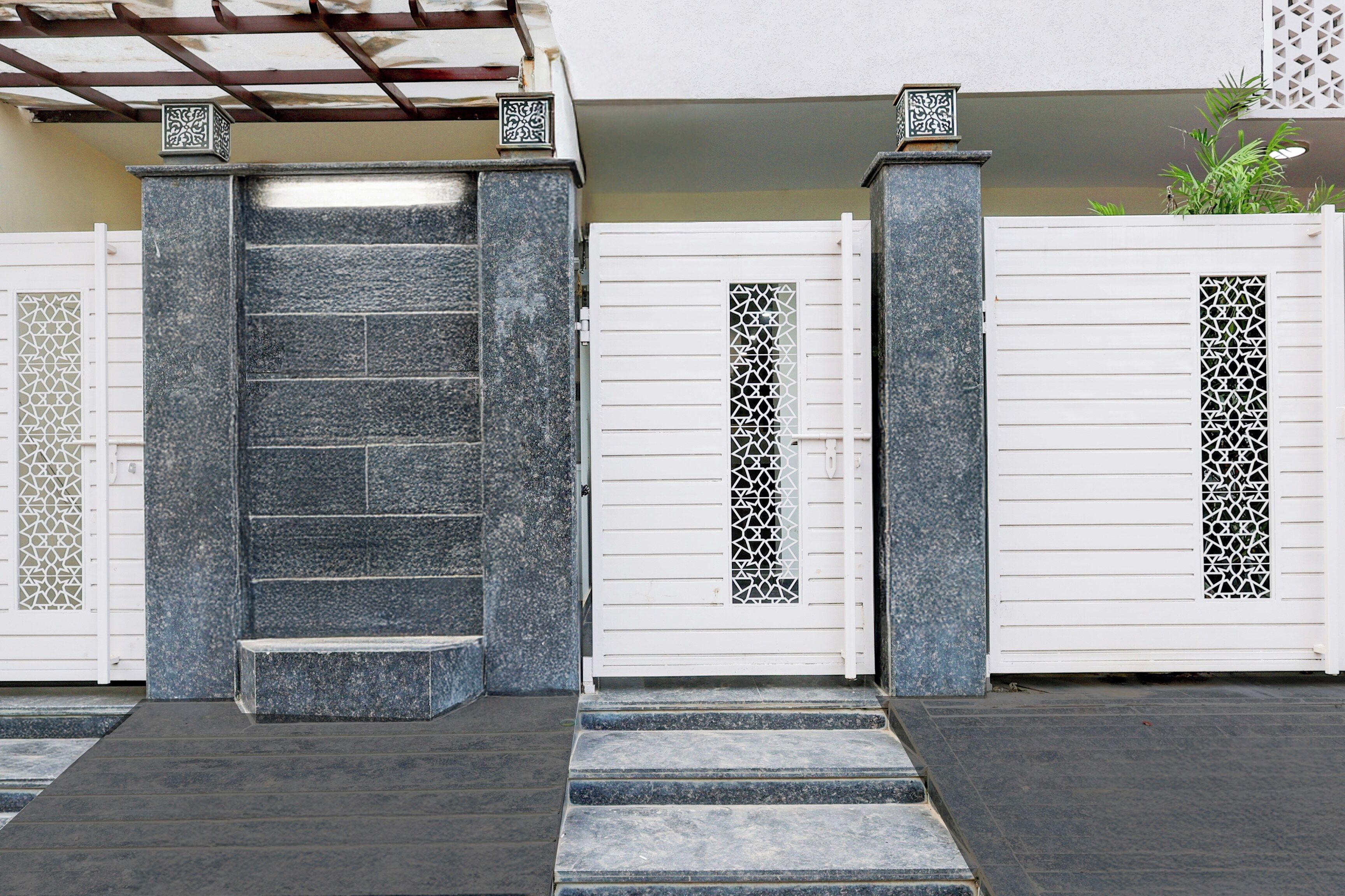 A gated entrance with a stone staircase leading up to a door, flanked by decorative pillars and a metal gate with intricate patterns.