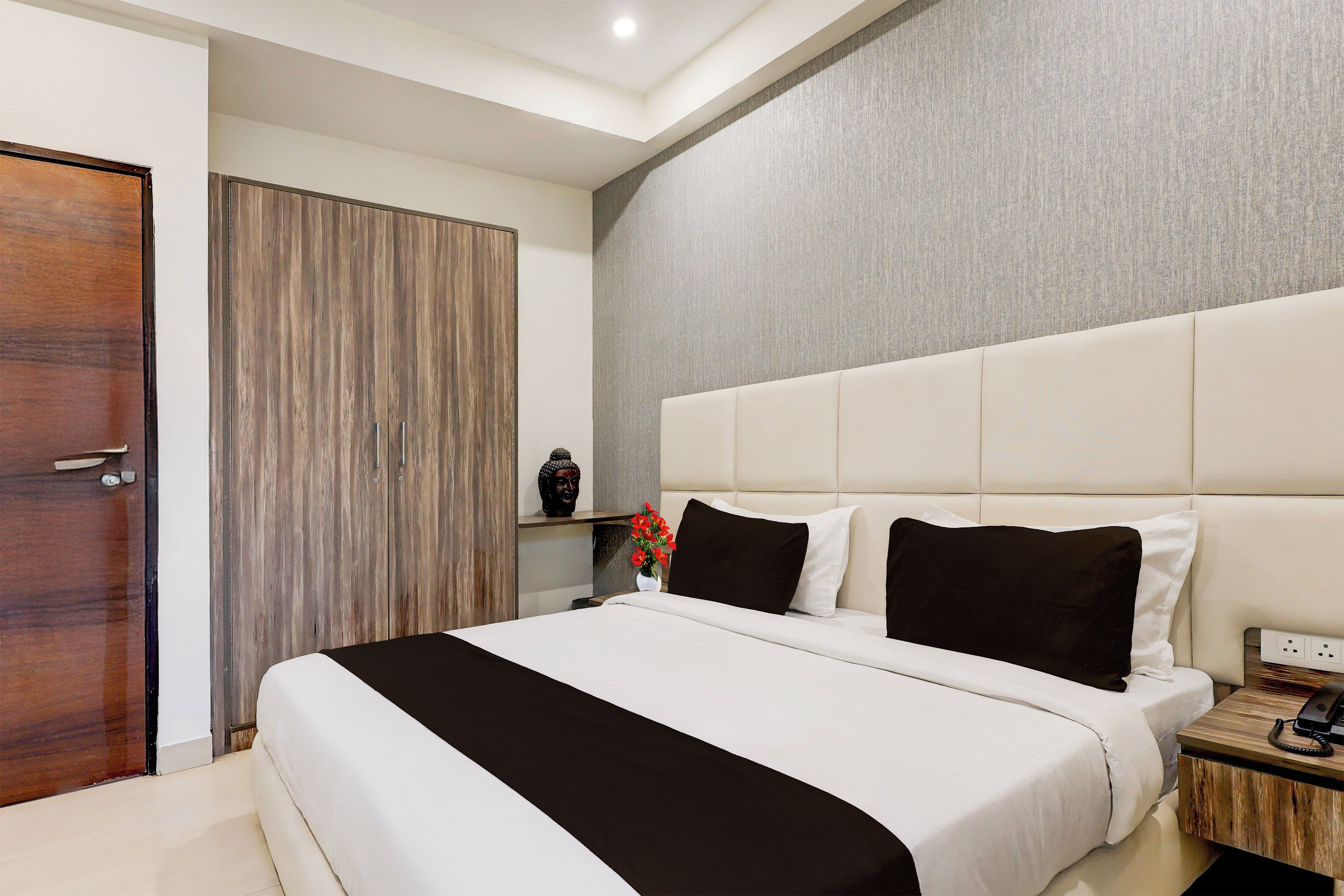 Een moderne slaapkamer met een groot bed, een nachtkastje met een telefoon en een wandplank met een Boeddhabeeldje en bloemen.