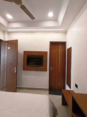 A hotel room with a bed, a television mounted on the wall, wooden doors, and a ceiling fan. - SRG Hotel and Restruant (Mathura)