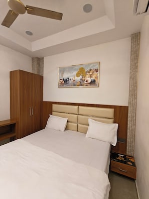A bedroom with a bed, a ceiling fan, a painting, a wardrobe, and a nightstand. - SRG Hotel and Restruant (Mathura)