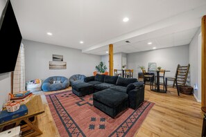 Una sala moderna con un sofá seccional, una mesa de centro y un tapete rojo estampado. Hay un comedor con una mesa y sillas de madera, y una escalera de madera apoyada en la pared.