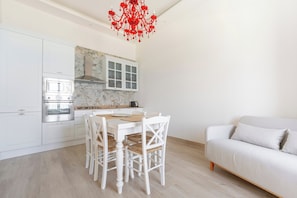 A modern kitchen with white cabinetry, a dining table with chairs, and a white sofa in the adjacent living area. - Ortigia Prestige (Siracusa)