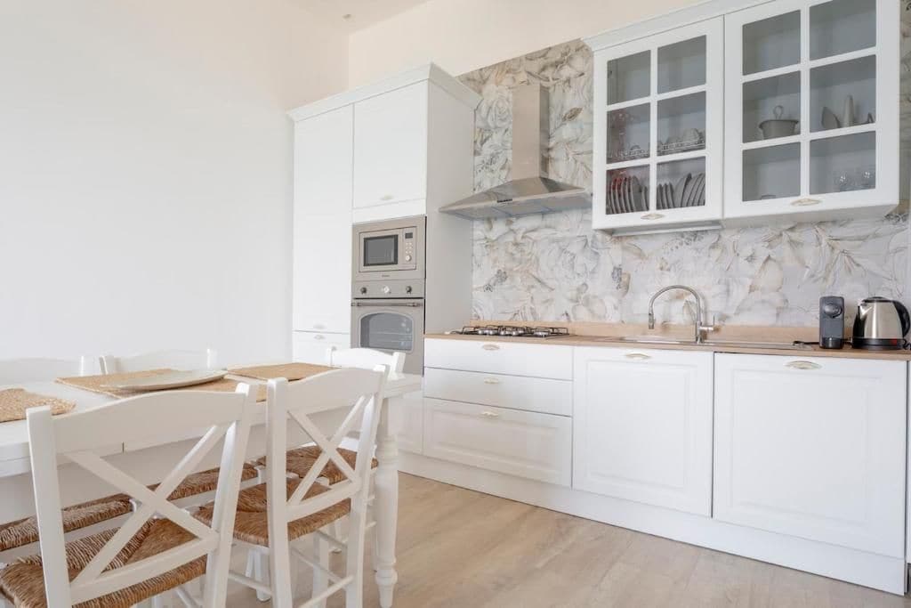 Una cocina moderna con armarios blancos, protector contra salpicaduras de mármol y un comedor con sillas y mesa blancos.
