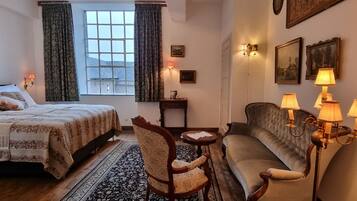 Een hotelkamer met een bed, een bank, een stoel, een bijzettafeltje, een raam met gordijnen, wandlampen en ingelijste foto's.